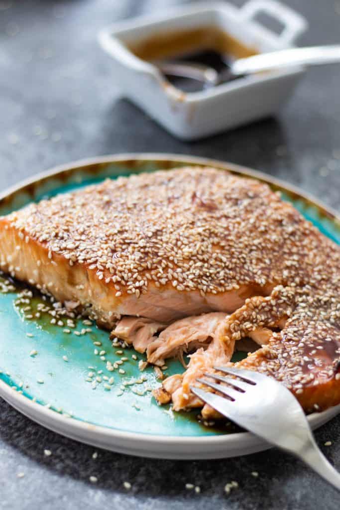 vue latérale du saumon teriyaki cuit au four sur une assiette turquoise avec une fourchette séparant le saumon, et un bol de sauce teriyaki en arrière-plan