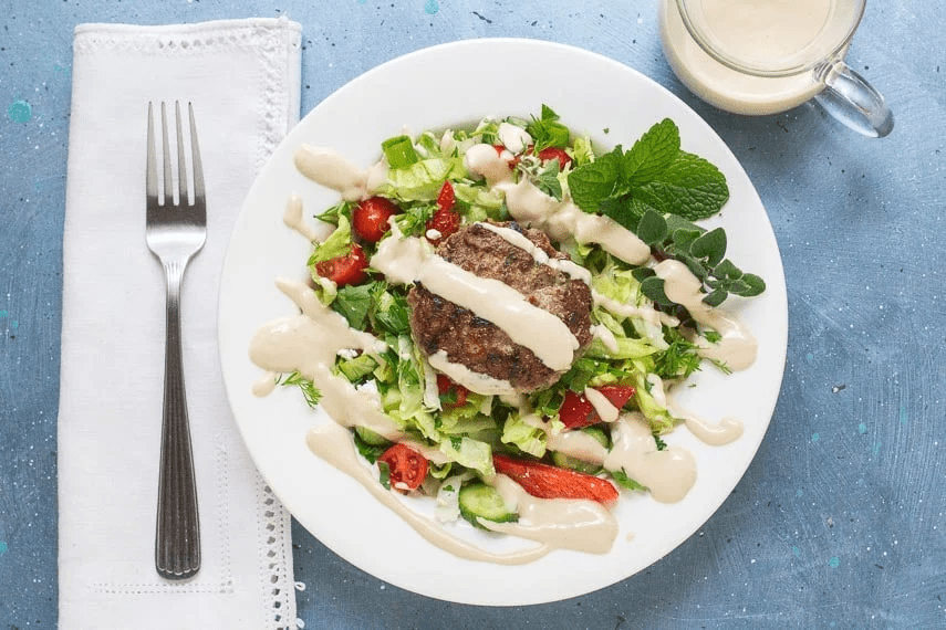 Une assiette avec salade, un hamburger d'agneau et une sauce.