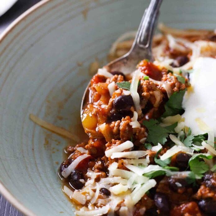 A green bowl with beef chili topped with sour cream, shredded cheese and fresh cilantro.